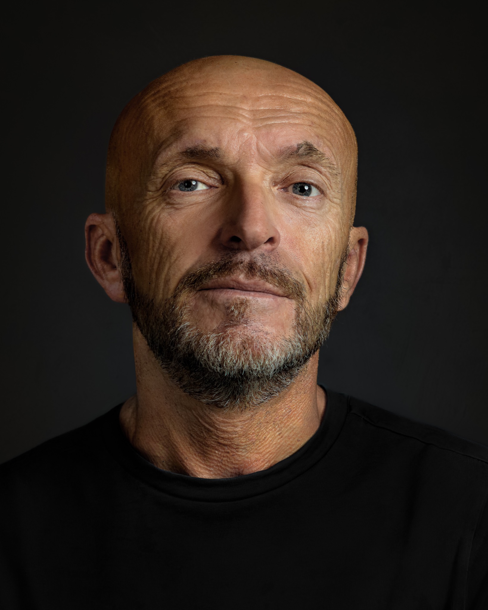 Headshot of a bald man with a short beard and a black shirt against a grey background