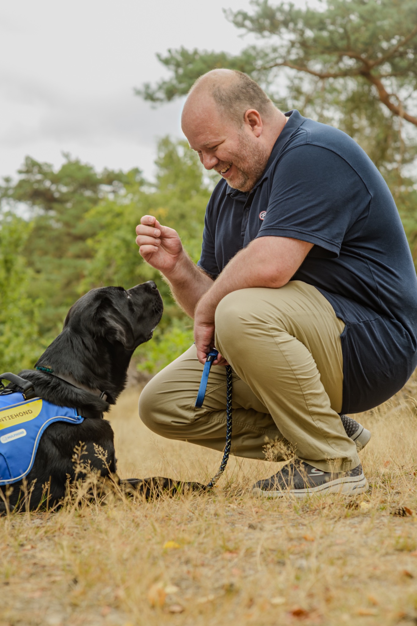 Een man knielt buiten naast een zwarte assistentiehond
