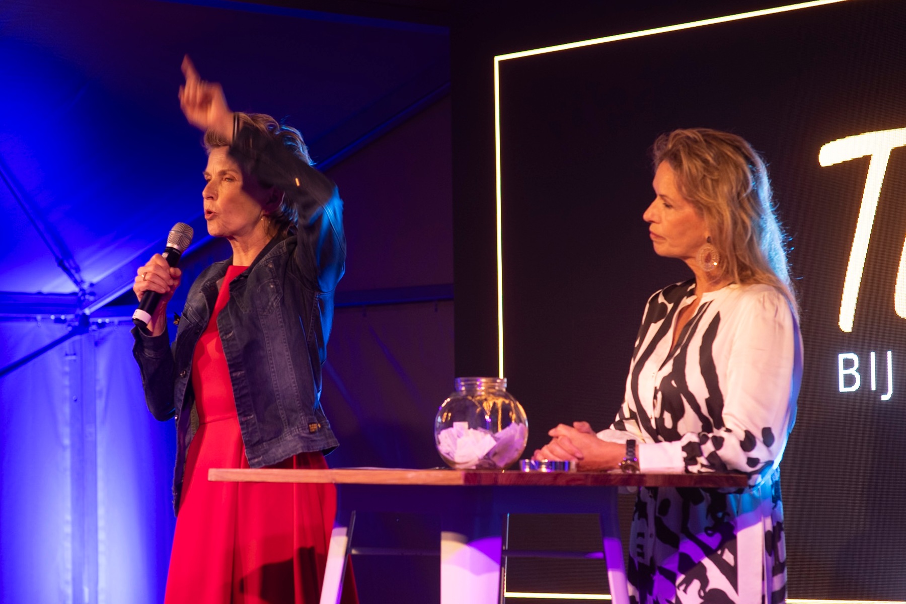 Two presenters on stage, one raising her hand beside the event backdrop.