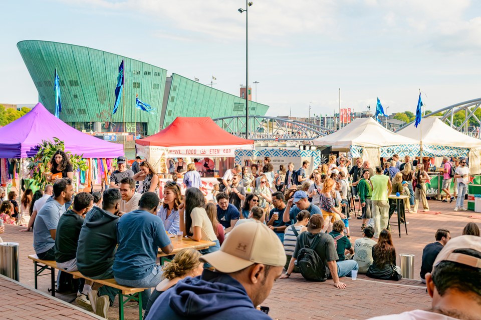 Guests gather at outdoor tables beside colourful food stalls, with NEMO across the water.
