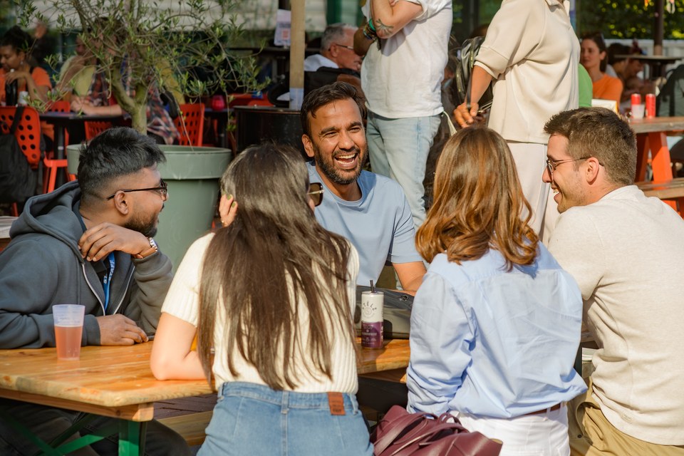 Friends laugh and talk around an outdoor wooden table.
