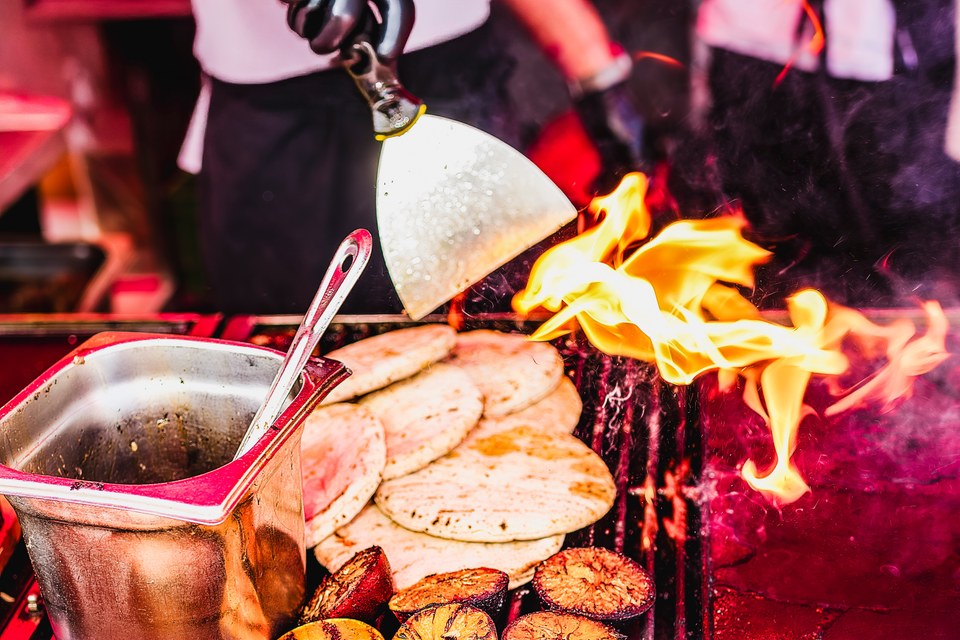 Flames rise beside flatbreads cooking on a grill.