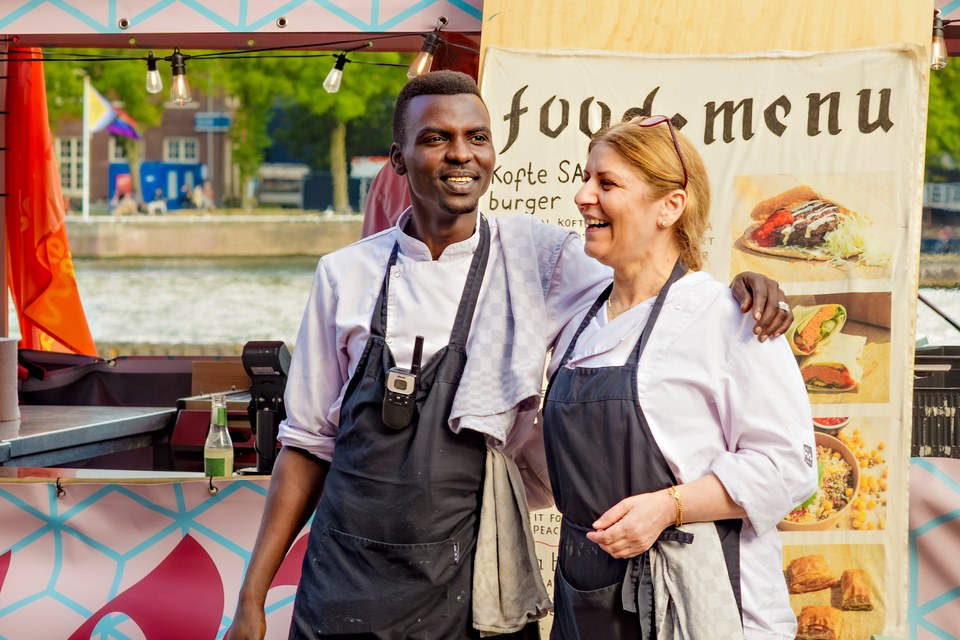 Two smiling cooks stand together in front of the food stall.