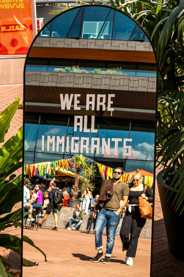 A mirror reflects visitors beneath the words We are all immigrants.