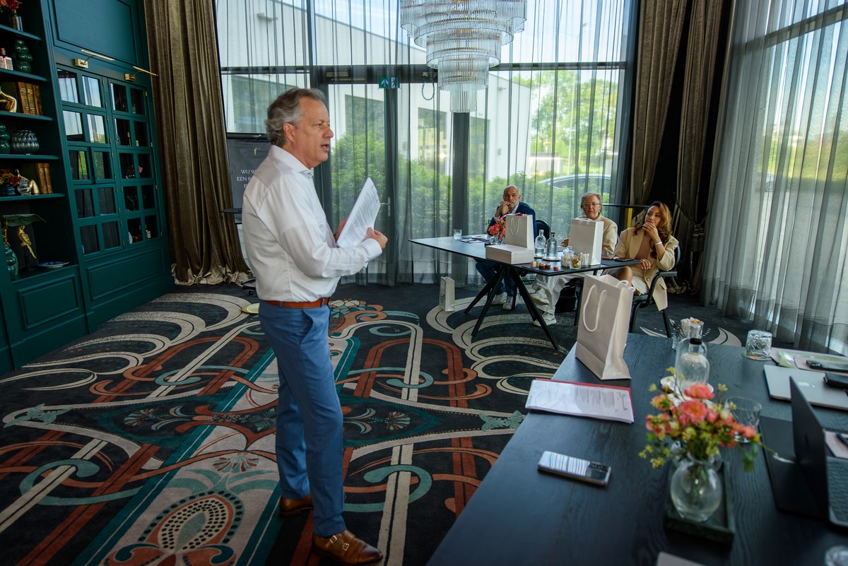 A man addresses the judging room beside two tables.
