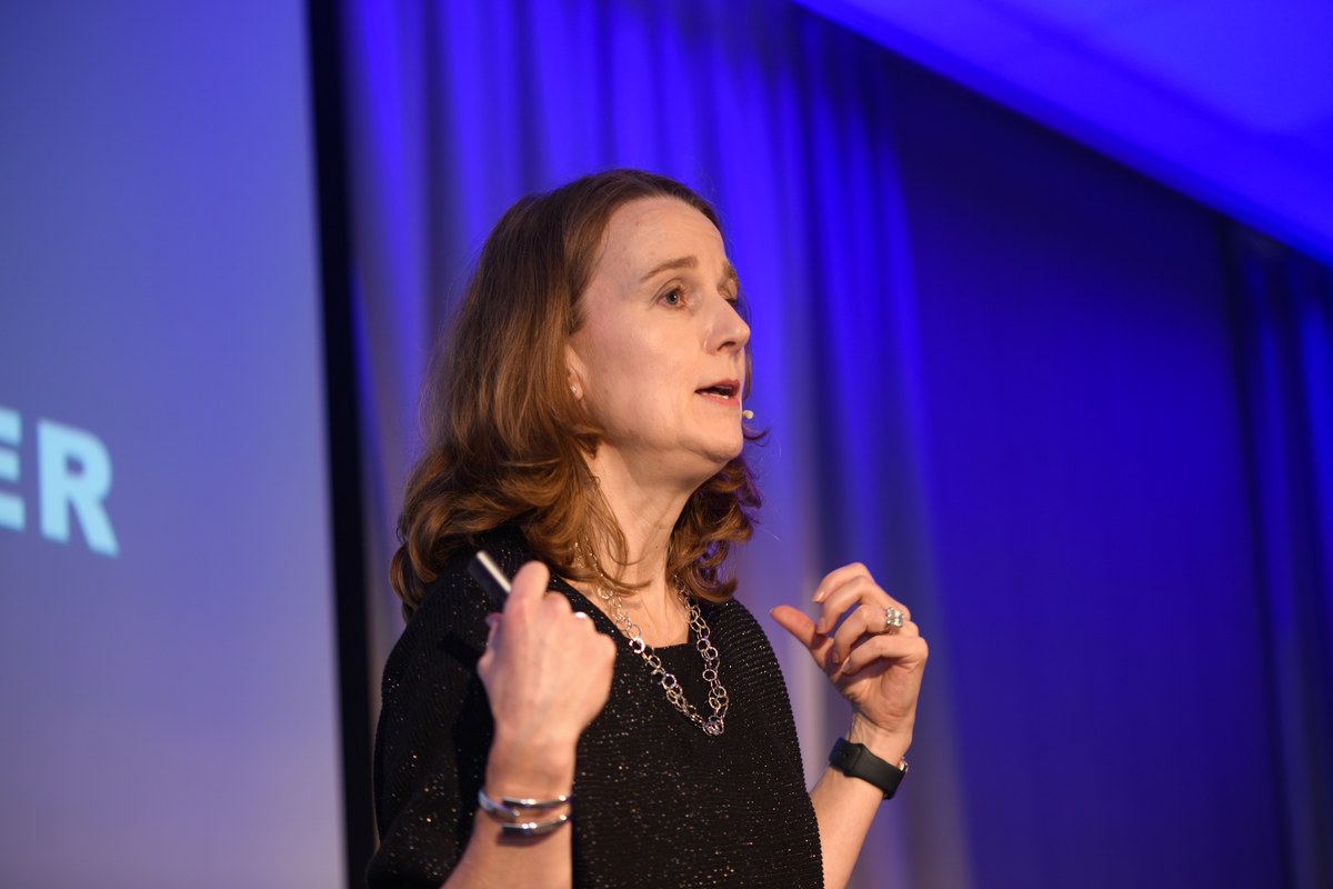A speaker gestures with both hands under blue stage lighting.