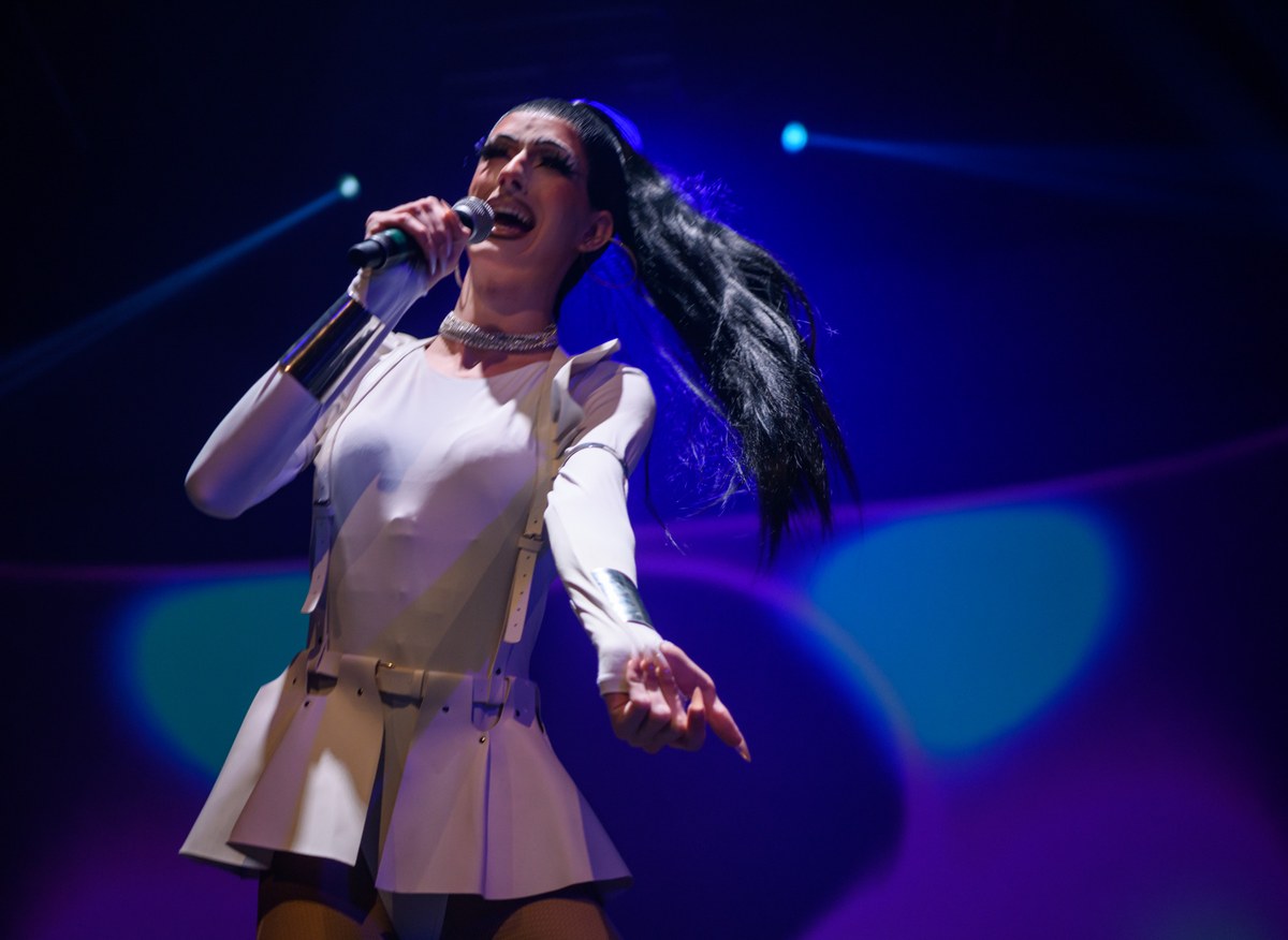 A performer in white holds a microphone as long dark hair swings across the blue-lit stage.