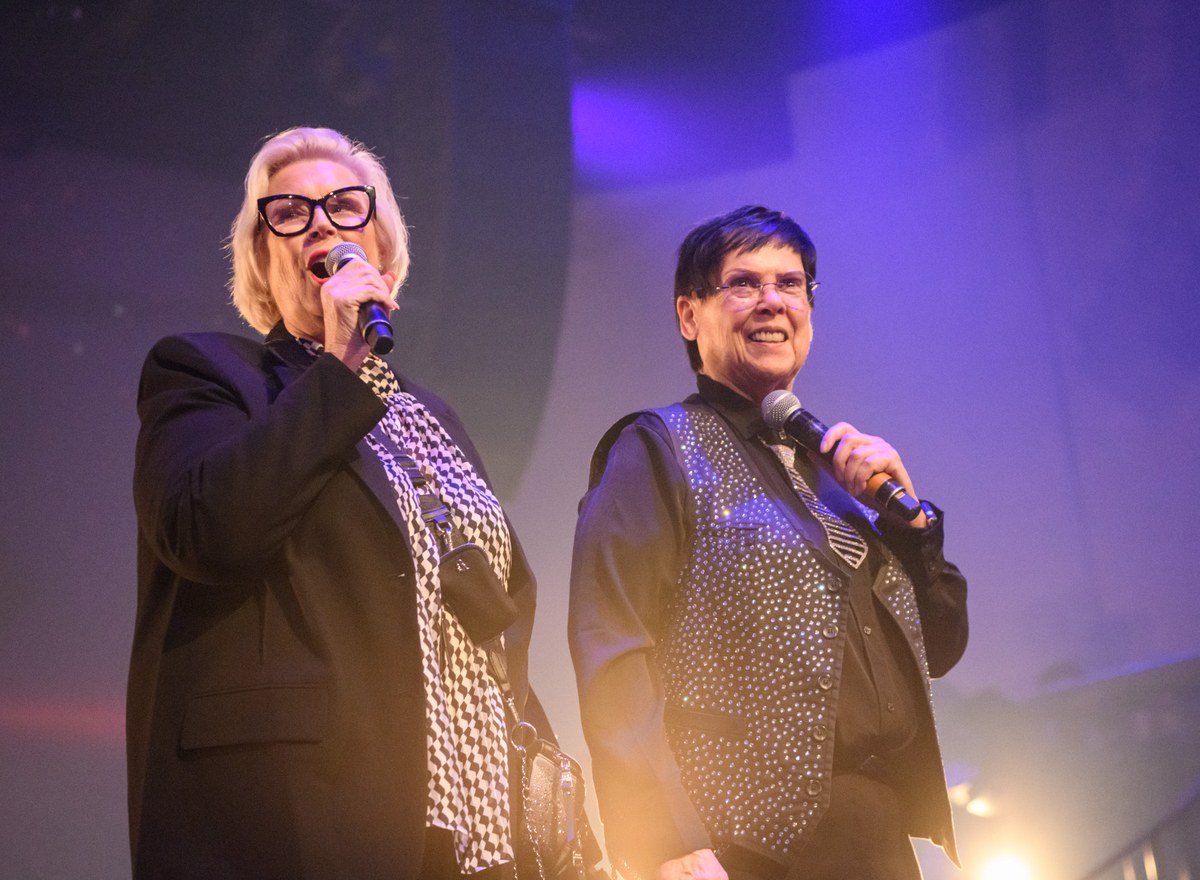 Two speakers hold microphones under purple stage lighting.