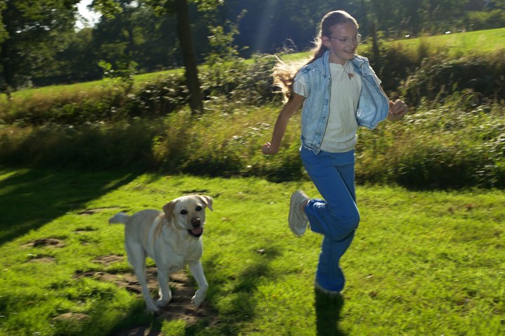 A girl runs through the grass beside an assistance dog.