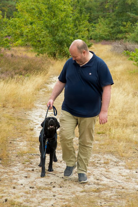 A man walks beside an assistance dog on a sandy track.
