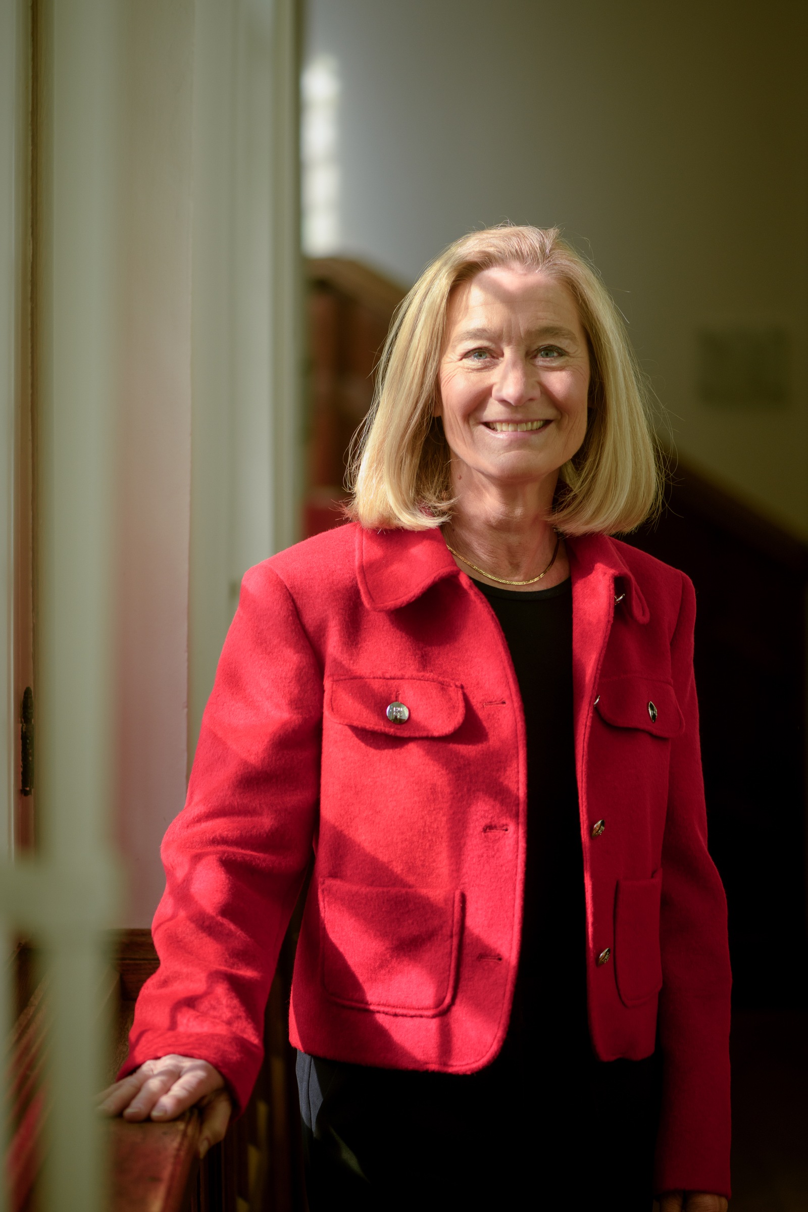 A woman smiling in a sunlit hallway, wearing a red jacket.