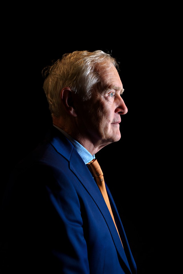 A man in profile against a dark background, in a blue suit and orange tie.