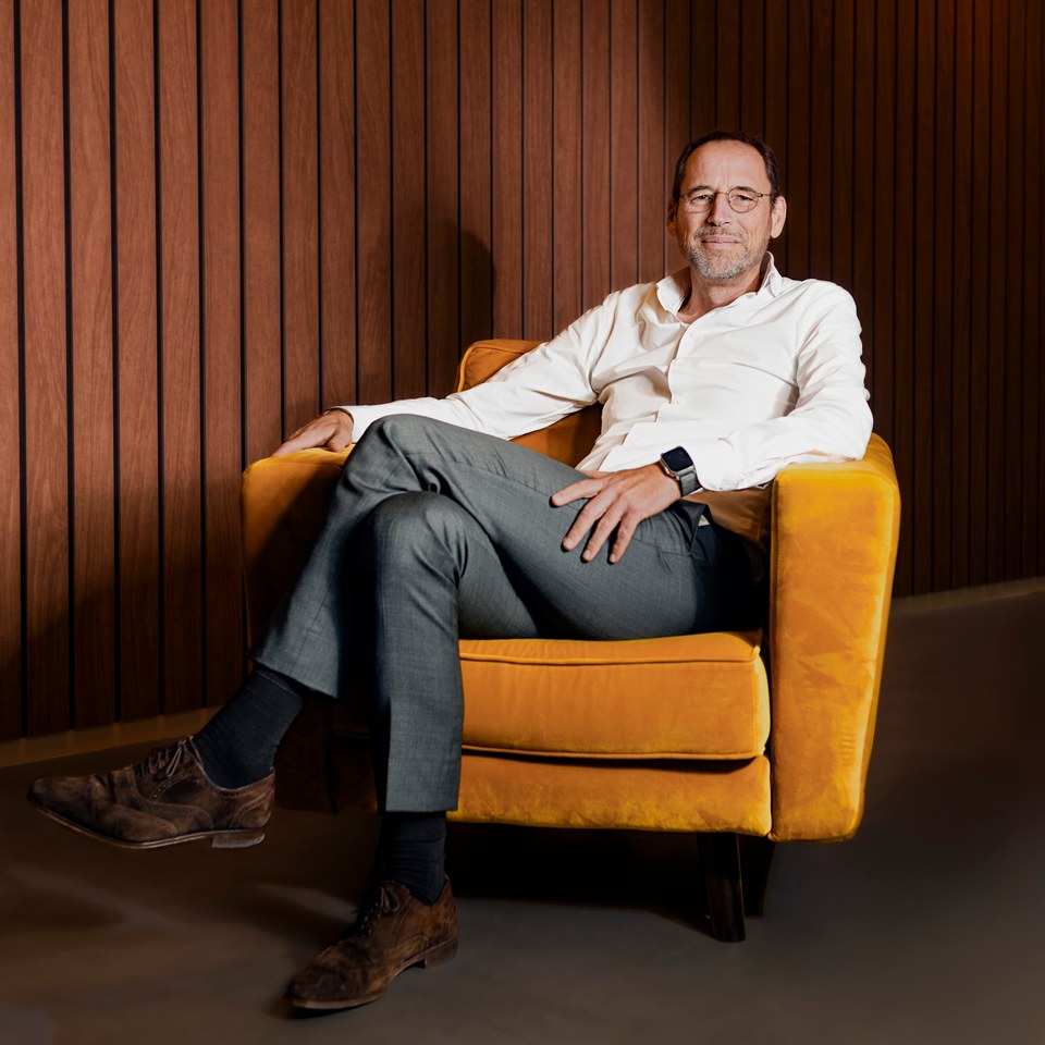 A man seated in a mustard-coloured chair against a wooden wall.