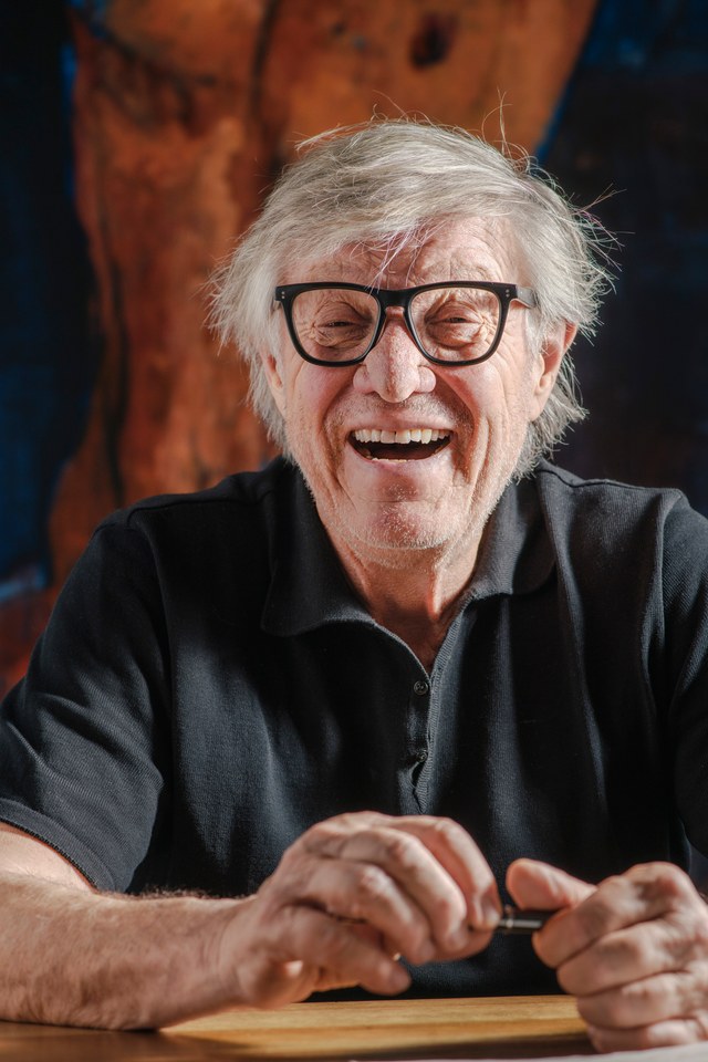 A man laughing at a wooden table in front of a painting.