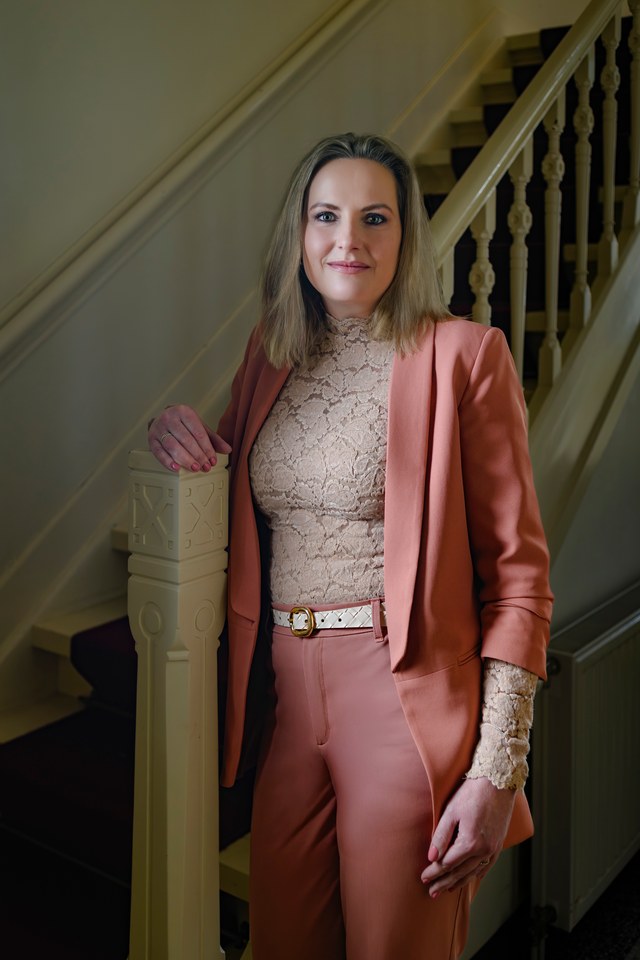 A woman in a salmon-coloured suit beside an ornate staircase.