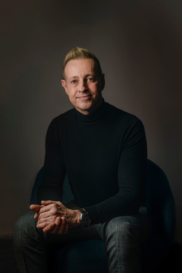 A man seated in a dark roll-neck sweater, hands together, against a plain dark background.