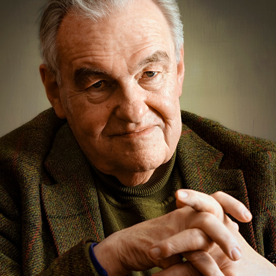 A man in a tweed jacket, hands clasped in the foreground and looking directly at the camera.