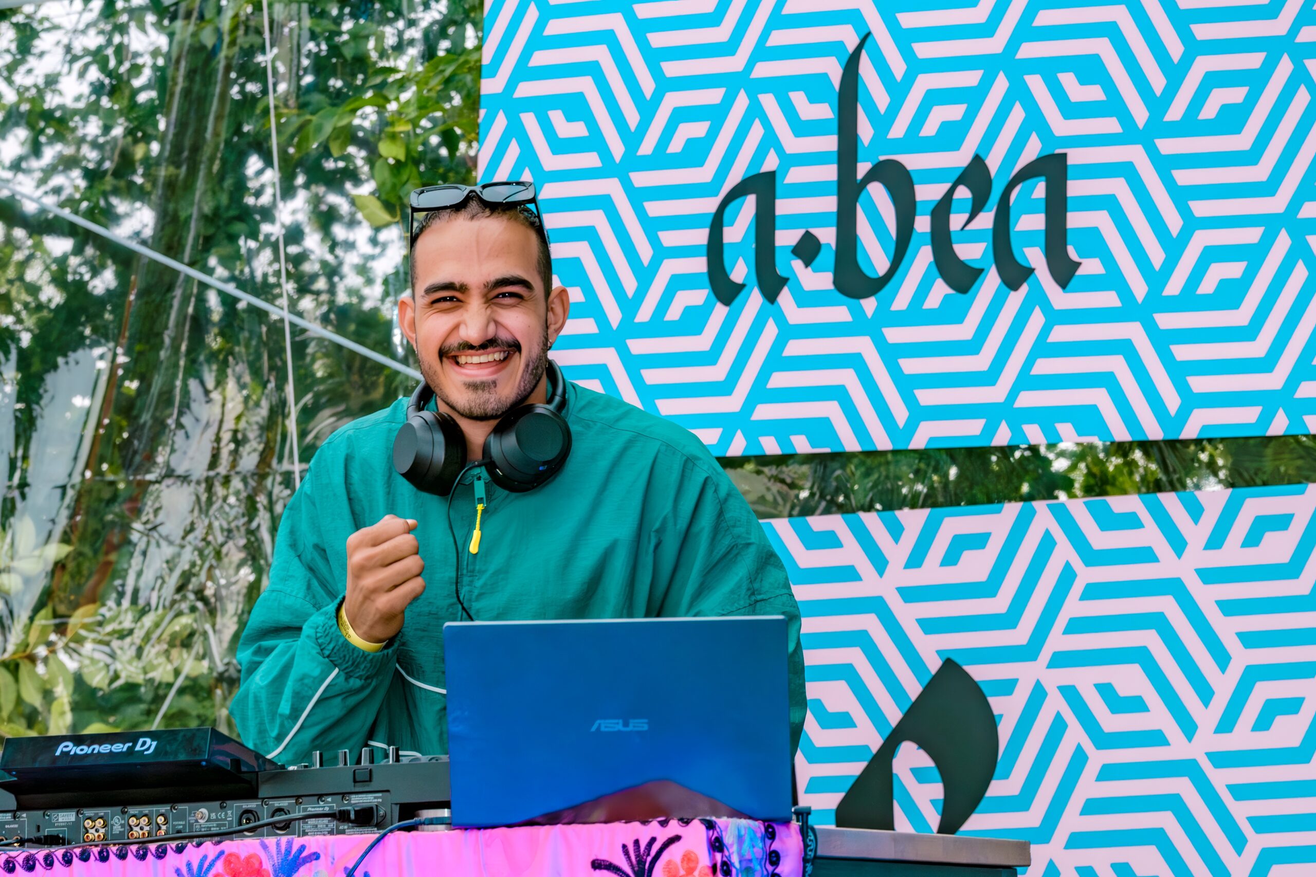 A smiling DJ in a green jacket with headphones around his neck behind his decks.