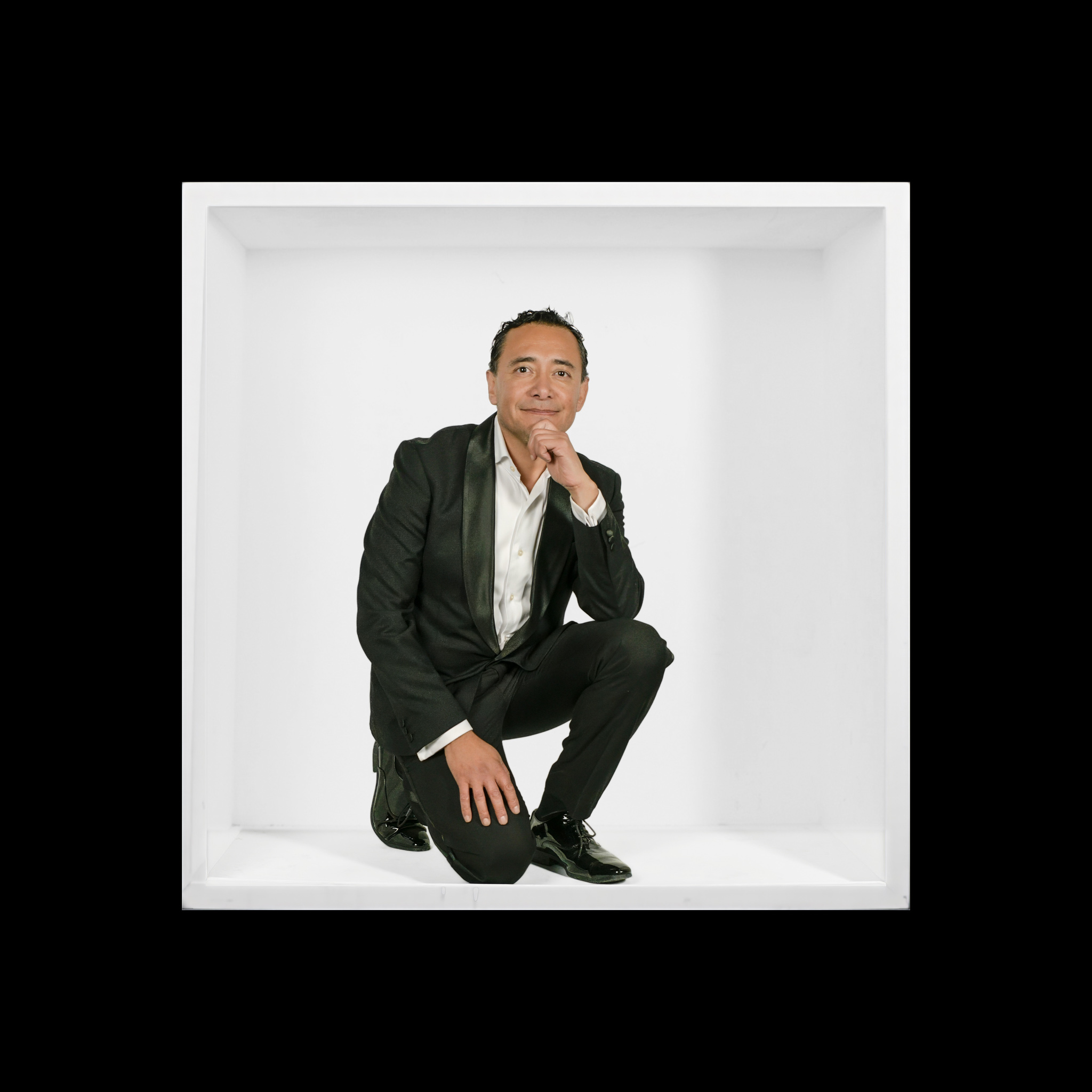Man in a dark suit kneeling in a white box, with his hand beneath his chin