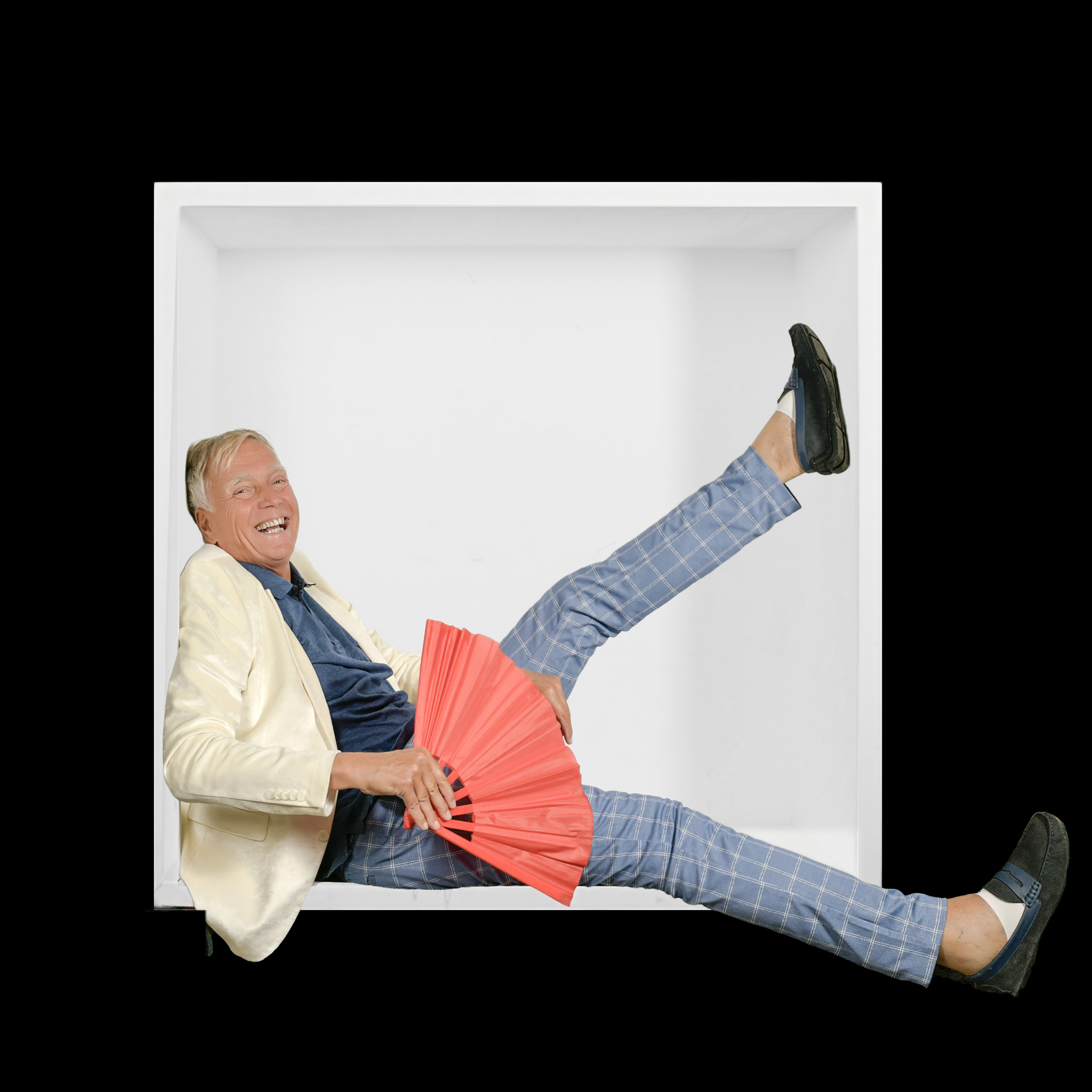 Laughing man in a cream jacket with a red fan, extending his legs beyond the white box