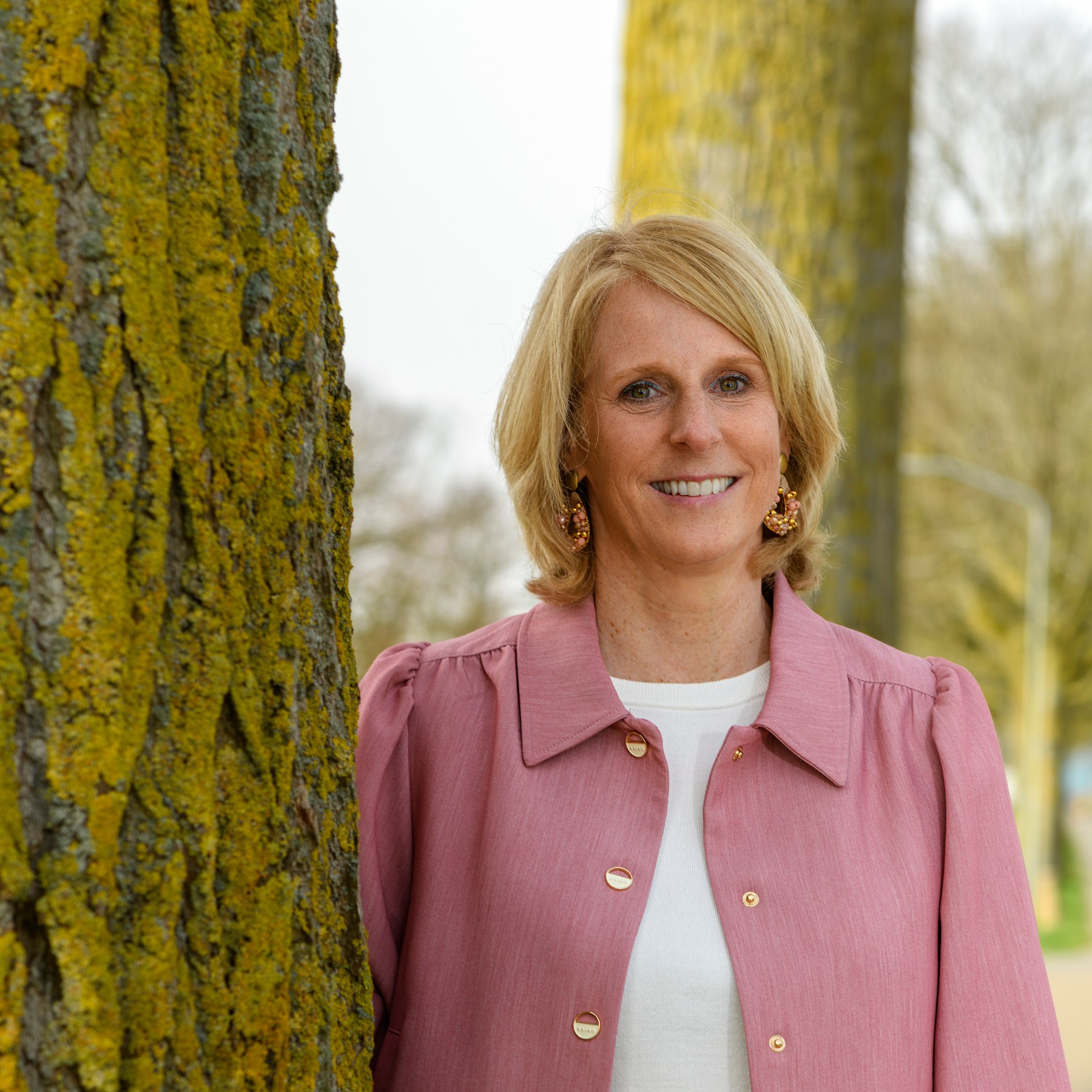 A smiling leader in a pink jacket beside a tree
