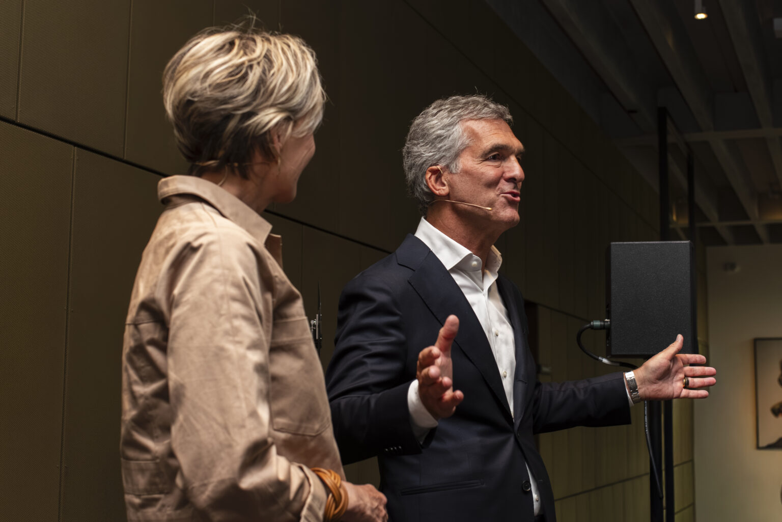 A speaker gestures with open arms beside the presenter during the anniversary programme.